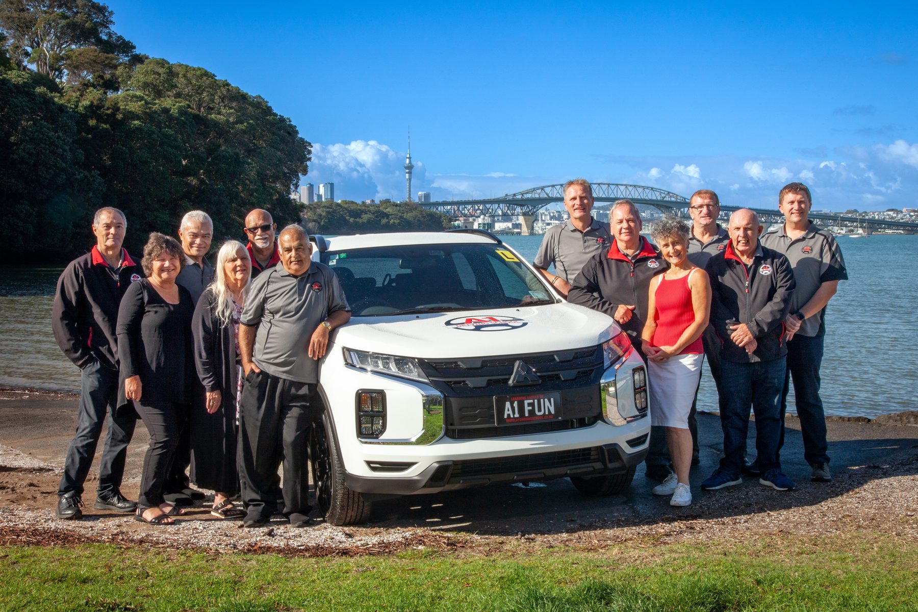 The A1 Driving School team beside their branded car, with the Auckland Harbour Bridge and Sky Tower behind